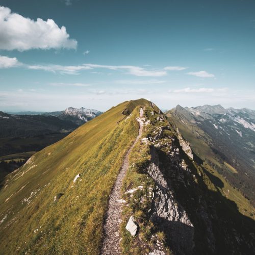 Sentier étroit longeant une crête montagneuse verdoyante, avec des sommets à l’horizon.